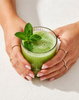 Hands holding a glass of green juice with mint leaves on a light background