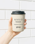 A hand holding a beige reusable coffee cup with a black lid, labeled 'KAILO Summit Experiences Curated by goop'.