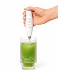 Hand using a KAILO frother to create foam in a glass of green liquid on a white background