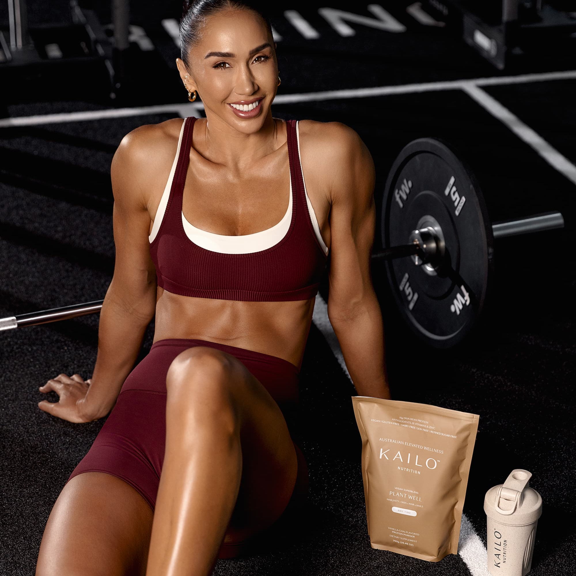 Woman in workout attire sitting on gym floor with protein powder and shaker.