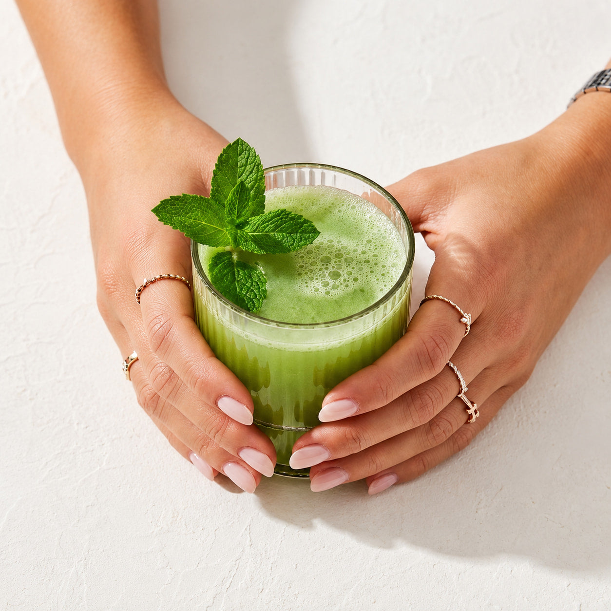 Hands holding a glass of green juice with mint leaves on a light background
