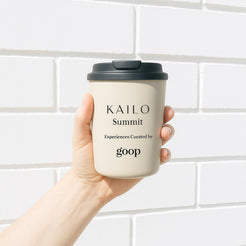 A hand holding a beige reusable coffee cup with a black lid, labeled 'KAILO Summit Experiences Curated by goop'.