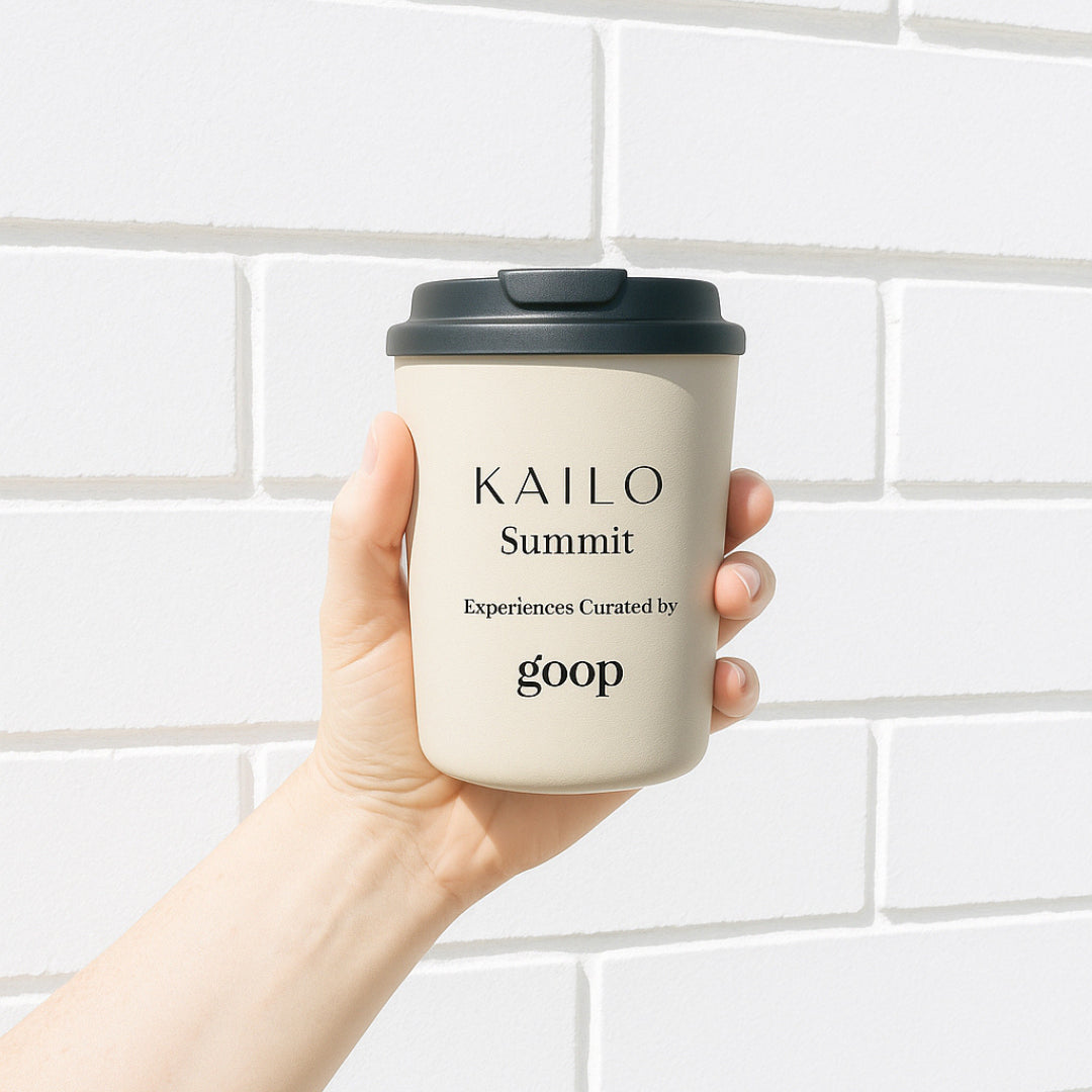 A hand holding a beige reusable coffee cup with a black lid, labeled 'KAILO Summit Experiences Curated by goop'.
