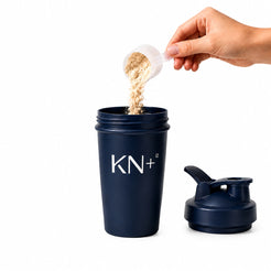 Navy blue shaker cup with 'KN+12' branding, being filled with powder from a white scoop.