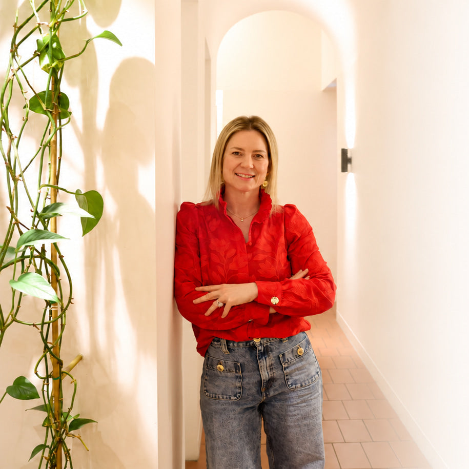 A woman with blonde hair wearing a red long-sleeve blouse and blue jeans stands with her arms crossed in a brightly lit hallway with white walls and a potted plant on the left.