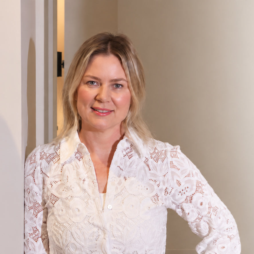 A blonde woman wearing a white lace long-sleeve blouse, standing in a doorway against a light beige wall.