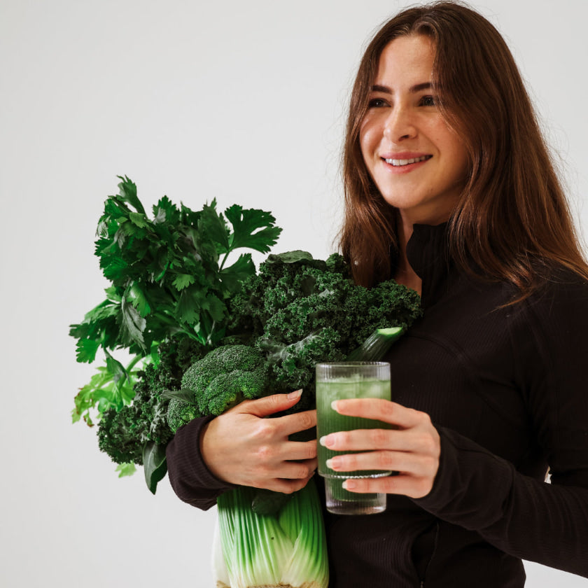 Woman holding a bunch of green vegetables and a glass of green juice against a white background