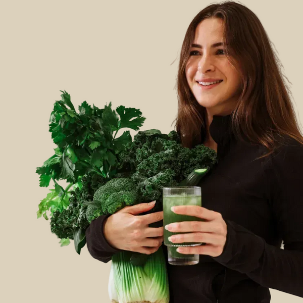 A smiling woman with long brown hair holds a large bunch of fresh green vegetables, including broccoli, kale, and parsley, next to a glass of green juice, against a plain light gray background.