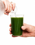 Person pouring green juice from a bottle into a glass on a white background