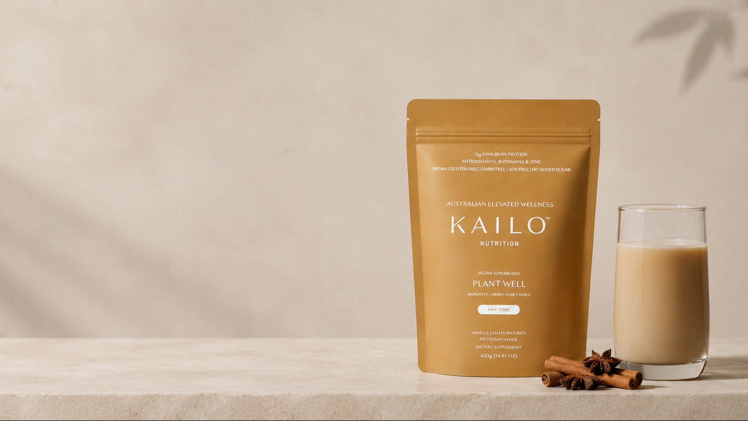 A stand-up pouch of KAILO Plant WELL supplement in a warm tan-gold color, standing on a light stone surface next to a glass of creamy beige drink and a few cinnamon sticks, against a softly textured beige wall with leaf shadows.