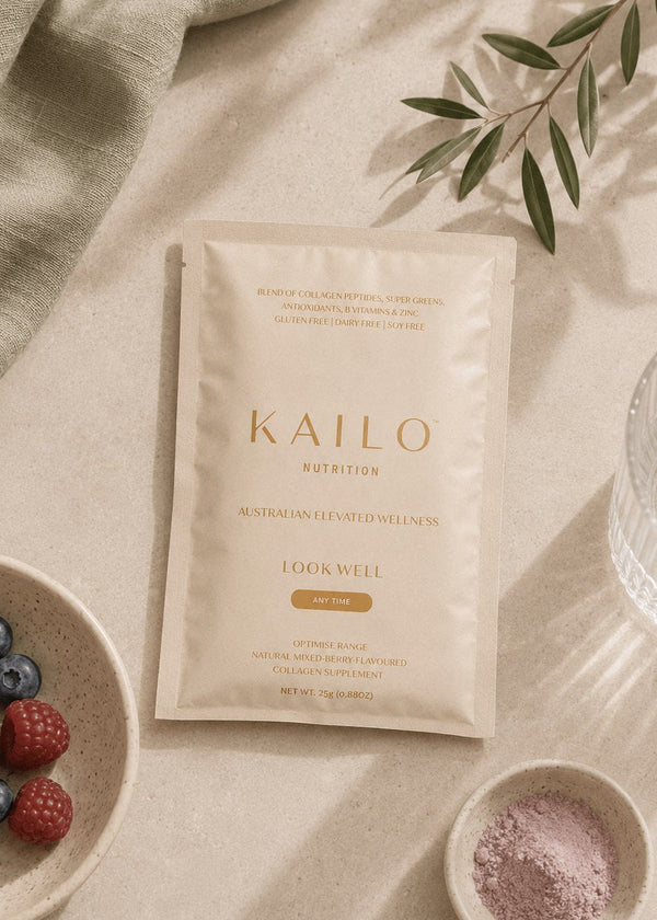 A Kailo Nutrition Lookwell skincare packet in a cream-colored stand-up pouch, styled in a flat-lay arrangement on a beige linen surface with a green leafy sprig, a bowl of berries, a small dish of pink powder, and a glass of water.