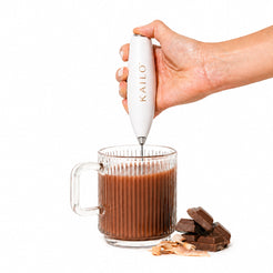Hand using a Kailo milk frother to stir a glass of hot chocolate with chocolate pieces and a spoon on a white background.