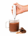 Hand using a Kailo milk frother to stir a glass of hot chocolate with chocolate pieces and a spoon on a white background.