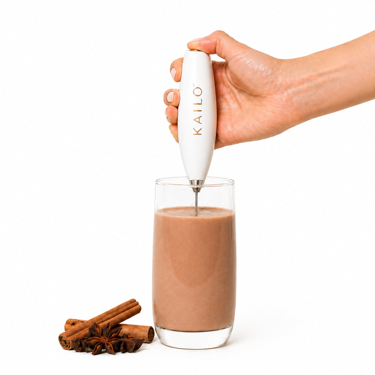 Hand using a Kailo whisk to froth a glass of chocolate milk with cinnamon sticks on a white background.